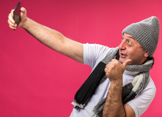 onder de indruk volwassen zieke blanke man met sjaal om de nek dragen winter hoed duimen omhoog kijken naar telefoon nemen selfie geïsoleerd op roze muur met kopie ruimte
