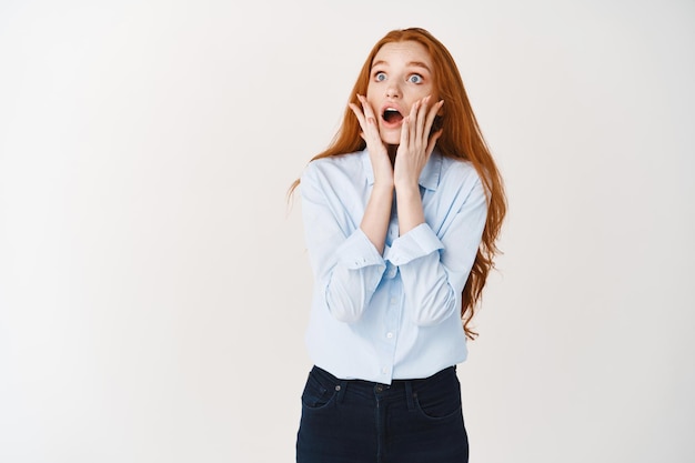 Onder de indruk vrouw met lang rood haar die promotiedeal bekijkt, staart naar linkerbovenhoek met geschokte uitdrukking, witte muur