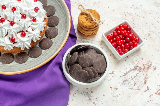 Onderaanzicht cake met banketbakkersroom op grijze plaat paarse sjaalkoekjes vastgebonden met touwbessen en chocolade in kom op witte tafel