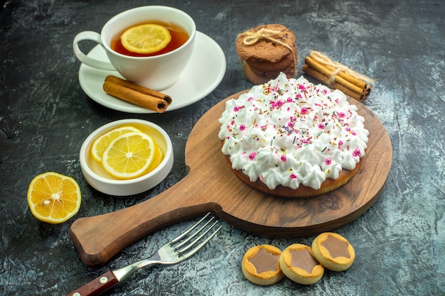 Onderaanzicht cake met banketbakkersroom op snijplank koekjes vork kaneelstokjes kopje thee op smaak gebracht met citroen en kaneel op grijze tafel