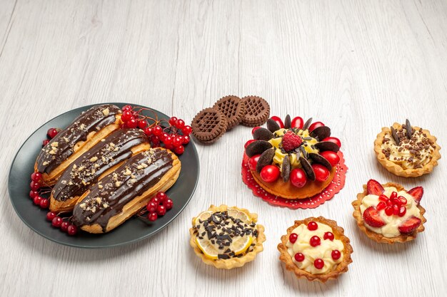 Onderaanzicht chocolade-eclairs en aalbessen op de grijze plaat, taartjes, koekjes en bessencake op de witte houten tafel met kopie ruimte