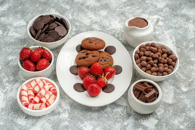 Onderaanzicht chocoladekoekjes aardbeien en ronde chocolaatjes op de witte ovale plaat en kommen met snoepjes aardbeien chocolaatjes granen en cacao op de grijs-witte achtergrond