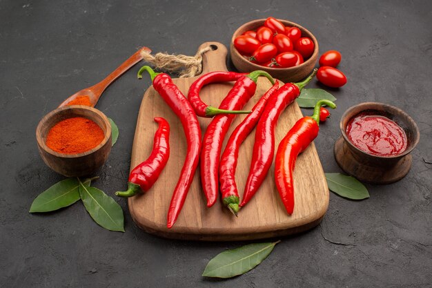 Onderaanzicht een kom met kerstomaatjes hete rode paprika's op de snijplank een houten lepel laurierblaadjes en kommen ketchup en hete peperpoeder op de zwarte tafel