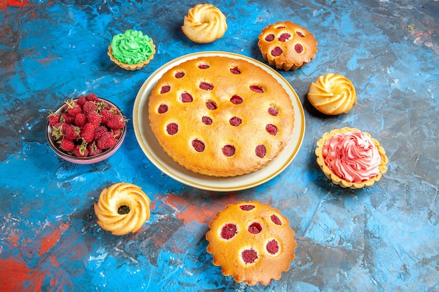 Onderaanzicht frambozencake op ovale plaat omringd door koekjes, kleine taartjeskom met frambozen op blauwe tafel