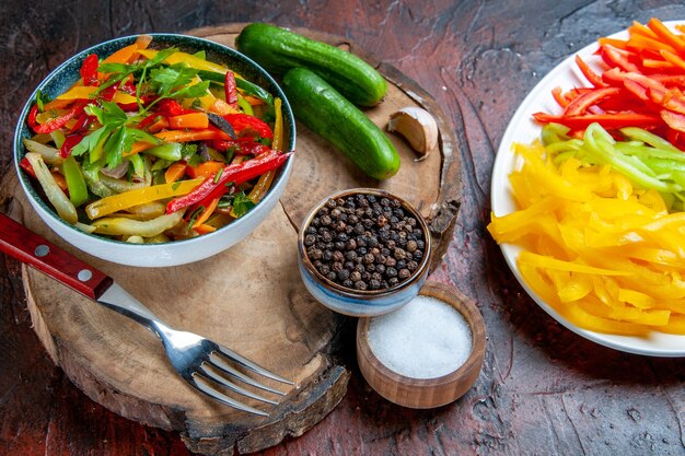 Onderaanzicht groentesalade in kom komkommers vork zwarte peper op rustiek bord zout gesneden pepers op donkerrode tafel