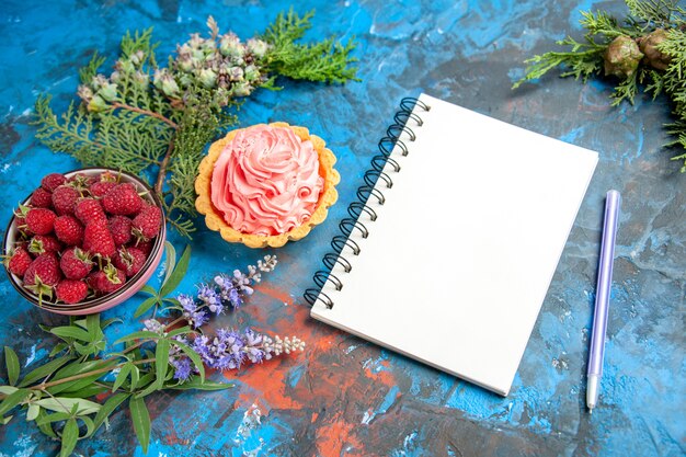 Onderaanzicht kleine taart met roze banketbakkersroomkom met frambozen een notitieboekje een pen op blauwe tafel