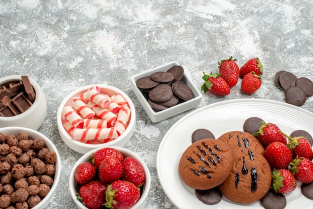 Onderkant sluiten koekjes aardbeien en ronde chocolaatjes op het ovale bord kommen met snoepjes aardbeien chocolaatjes granen op de grijs-witte tafel
