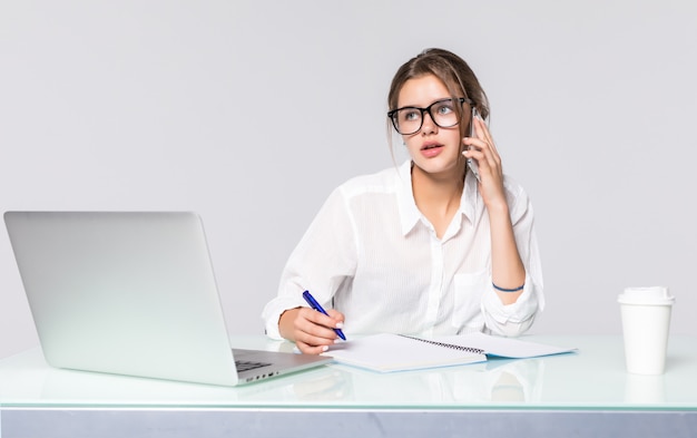 Onderneemster bij haar bureau met laptop en sprekende telefoon die op witte achtergrond wordt geïsoleerd