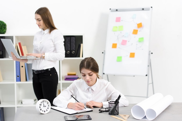 Onderneemster het schrijven nota&#39;s over klembord met haar vrouwelijke collega die digitale tablet in het bureau gebruiken