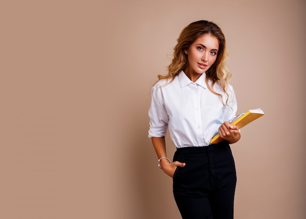 Onderneemster in modieuze oogglazen die boeken houden en zich over beige muur bevinden. Blond golvende haren. Witte blouse en zwarte broek.