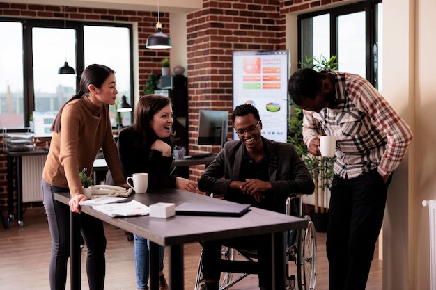 Ondernemer zit in een rolstoel en praat met zakenpartners tijdens werkpauze, lacht en heeft plezier in een gehandicaptenvriendelijk kantoor. Mannelijke werknemer met een handicap chatten met collega's.