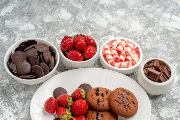 Onderste helft weergave koekjes aardbeien en ronde chocolaatjes op de witte ovale plaat omgeven kommen met snoepjes aardbeien en chocolaatjes op de achtergrond