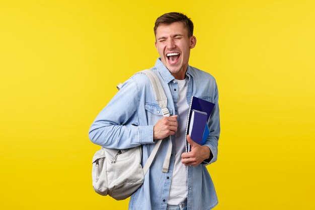 Onderwijs, cursussen en universitair concept. Vrolijke gelukkige knappe mannelijke student die geniet van het leven op de campus, rugzak en studiemateriaal vasthoudt, lachend en glimlachend opgewonden, gele achtergrond.