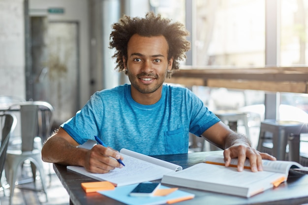 Onderwijs en kennis, Mensen en levensstijl. Indoor portret van vrolijke donkerhuidige student die thuisopdracht doet over wiskunde, werkt in een coffeeshop, notities maakt uit het leerboek