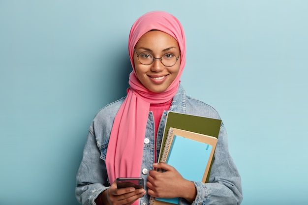 Onderwijs en religie concept. Spirituele gemengd ras vrouwelijke student gebruikt mobiele telefoon voor surfen op internet, houdt noodzakelijke notitieboekjes om te schrijven, draagt ronde bril en roze sluier, staat binnen