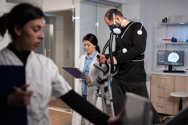 Onderzoeker arts kijkt naar monitor die de hartslag analyseert terwijl sportman draait op gymtrainer die aan lichaamsuithoudingsvermogen werkt tijdens medische training in modern lab. arts controleert ecg-gegevens