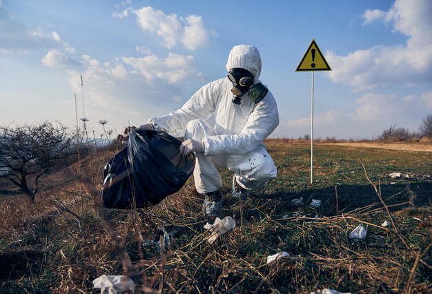 Onderzoeker in beschermend pak die op zonnige dag buiten plastic afval in zwarte afvalzak verzamelt