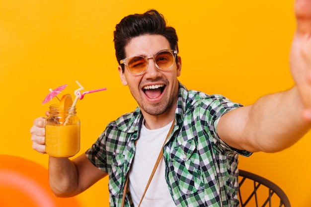 Gratis foto ondeugende brunette man in oranje zonnebril neemt selfie, knipoogt en houdt zoete cocktail.