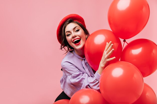 Ondeugende vrouw in rode baret lacht en heeft plezier op roze achtergrond met grote ballonnen.