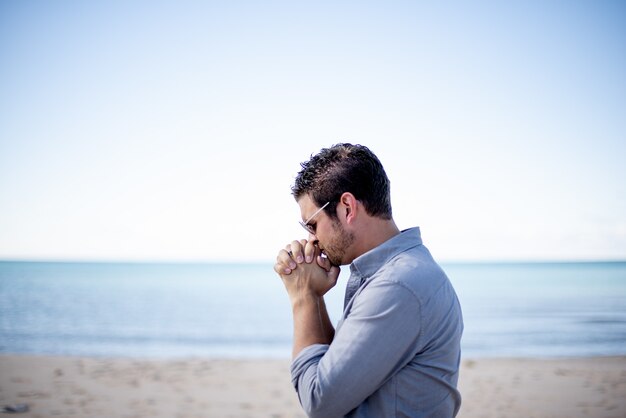 Ondiepe focus shot van een man in de buurt van het strand met zijn handen in de buurt van zijn mond tijdens het bidden