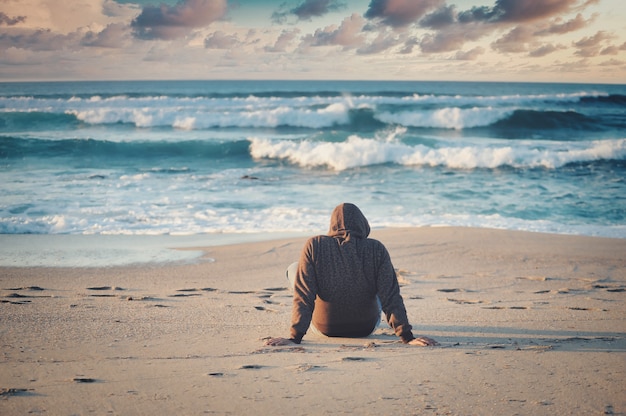 Ondiepe focus shot van een man in een zwarte jas zittend op een zandstrand