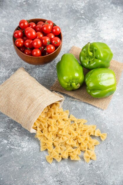 Ongekookte pasta's in een rustieke zak met kerstomaatjes en paprika's op marmeren tafel.