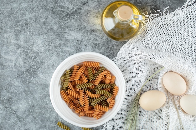 Gratis foto ongekookte spiraalvormige macaroni in witte plaat met drie eieren en een fles olie