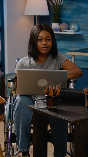 Ongeldige zwarte persoon met laptopcomputer die kunstwerken in creatieve ruimte thuis ontwerpt. Artistieke Afro-Amerikaanse vrouw in rolstoel die met apparaat werkt op zoek naar inspiratie voor meesterwerk