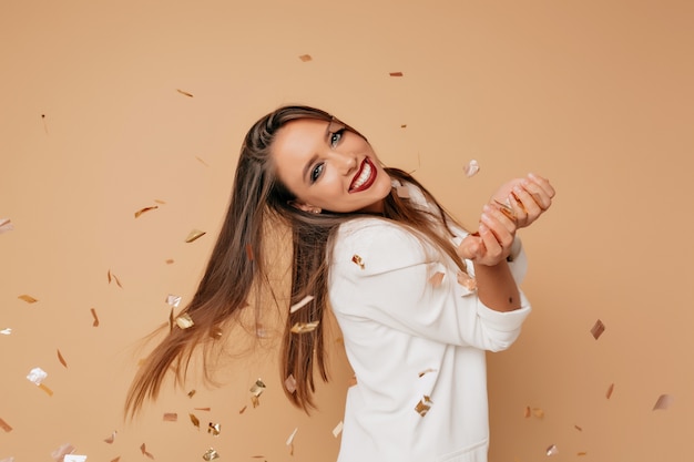 Ongelooflijk vrouwelijk model met mooie glimlach en lang lichtbruin haar dragen witte jas poseren op beige muur met confeti en verjaardagsfeestje voorbereiden