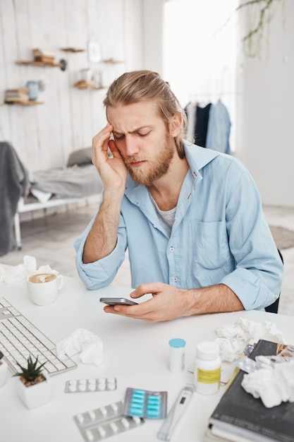 Ongelukkig bebaarde jonge mannelijke manager aan tafel zitten omringd door pillen, tabletten, drugs. Blonde kantoormedewerker is erg verkouden, surft op internet, heeft last van hoge temperaturen. Gezondheidsproblemen.