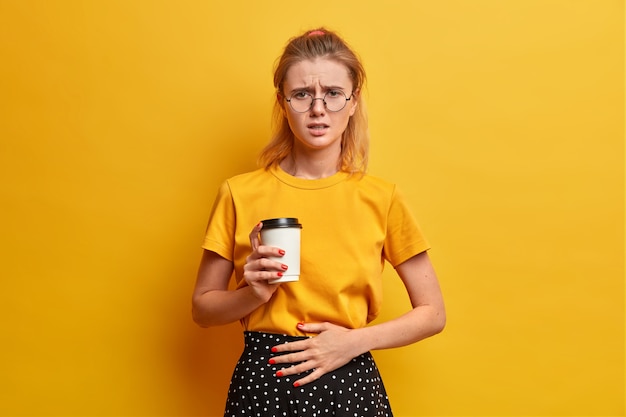 Ongelukkig ontevreden vrouw fronst gezicht, voelt zich onwel, houdt de hand op de buik, drinkt afhaalkoffie, at bedorven voedsel, draagt een transparante bril, casual geel t-shirt