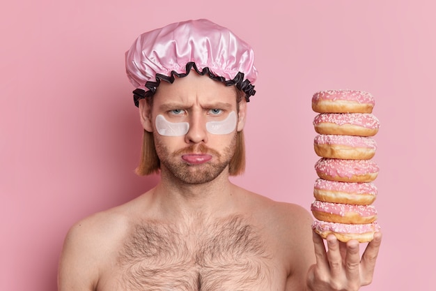 Ongelukkige man met sombere uitdrukking staat topless binnen houdt stapel zoete donuts past collageenpleisters toe om rimpels onder de ogen te verminderen.