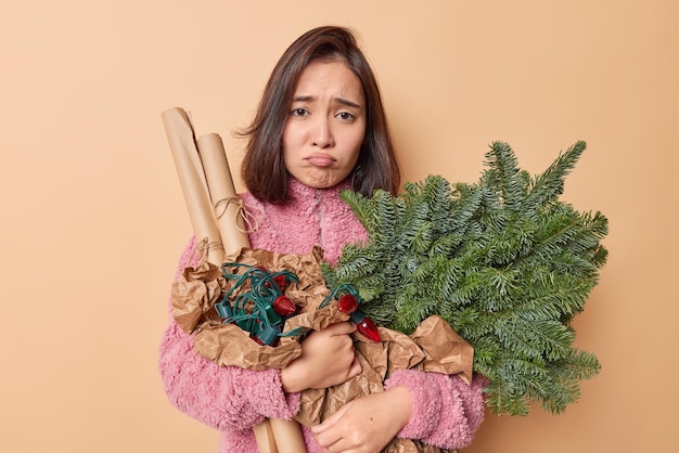 Ongelukkige sombere brunette vrouw met oosterse uitstraling houdt vakantie artikelen voor decoratie bereidt zich voor op nieuwjaarsviering draagt winterjas geïsoleerd over beige achtergrond. Kerstavond concept