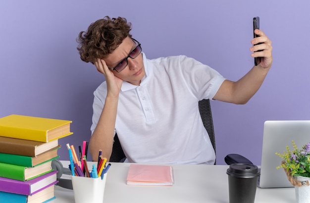 Ongelukkige student in een wit poloshirt met een bril die aan de tafel zit met boeken die selfie maken met een smartphone die ontevreden over een blauwe achtergrond kijkt