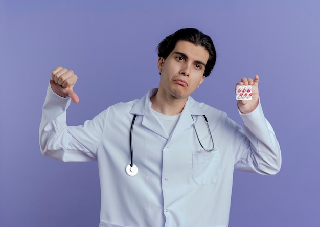 Ongenoegen jonge mannelijke arts die medische mantel en stethoscoop draagt die naar kant kijkt die pak medische capsules en duim omlaag toont geïsoleerd op paarse muur