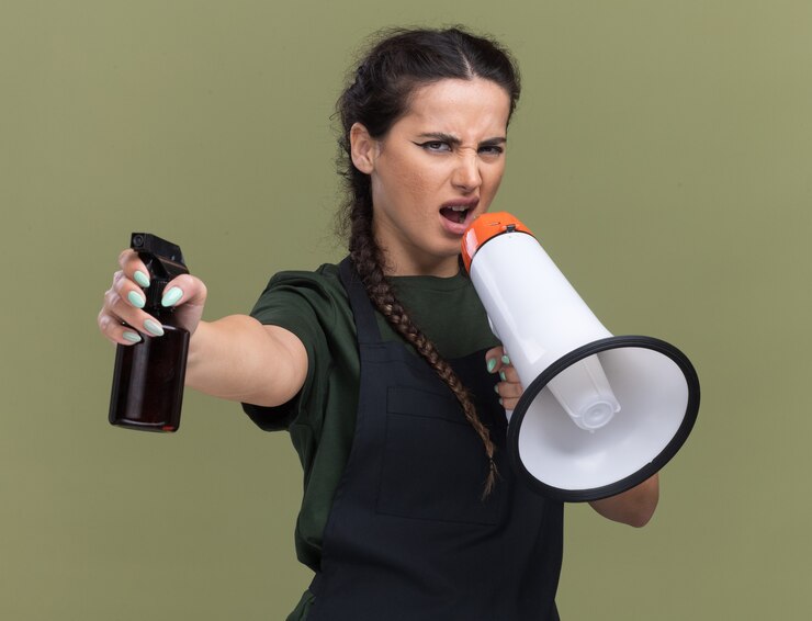 Gratis Foto ongenoegen jonge vrouwelijke kapper in uniform spreekt op luidspreker met spuitfles op camera geïsoleerd op olijfgroene muur
