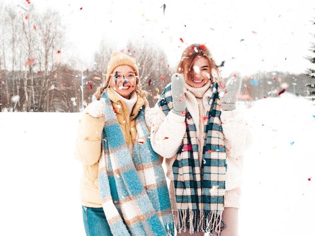 Ongericht twee jonge mooie lachende hipster vrouw in trendy warme kleding en sjaals. vrouwen poseren in de straat in het park. positieve pure modellen die plezier hebben in de sneeuw. genieten van winterse momenten. confetti