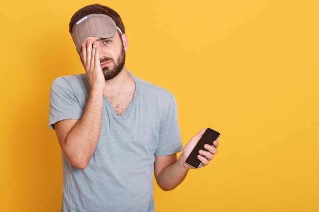 Ongeschoren donkerharige man kleedt slaapmasker, houdt smartphone in de hand en bedekt de helft van zijn gezicht met de handpalm, ziet er moe uit