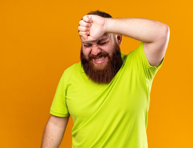 Ongezonde bebaarde man in geel poloshirt voelt zich vreselijk en lijdt aan kou die zijn voorhoofd aanraakt en lijdt aan sterke hoofdpijn die pijn voelt