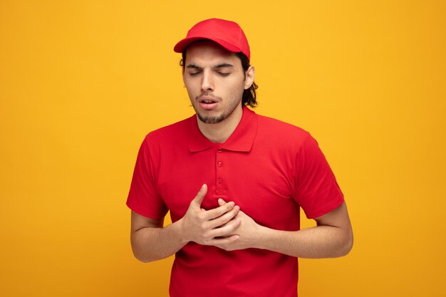 ongezonde jonge bezorger met uniform en pet die handen op de borst legt terwijl hij zijn ogen gesloten houdt geïsoleerd op gele achtergrond