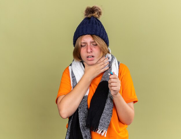 Ongezonde jonge vrouw die hoed met sjaal om haar nek draagt en zich onwel voelt terwijl ze verschillende pillen vasthoudt die lijden aan keelpijn wat betreft pijn in de nek die zich over de groene muur bevindt