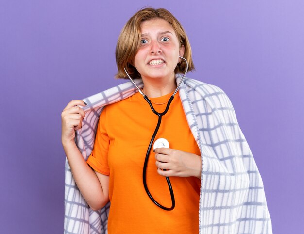 Ongezonde jonge vrouw gewikkeld in een warme deken die zich ziek voelt terwijl ze naar haar hartslag luistert met een stethoscoop en er bezorgd uitziet terwijl ze over een paarse muur staat