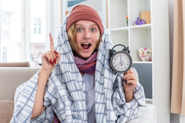 Ongezonde jonge vrouw in hoed gewikkeld in deken met wekker verrast tonend wijsvinger met goed idee zich beter voelen zittend op de bank in lichte woonkamer