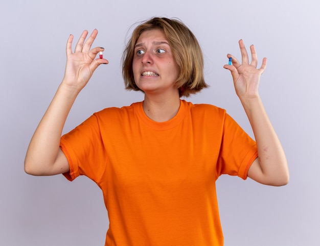 Ongezonde jonge vrouw in oranje t-shirt die zich onwel voelt en lijdt aan griep met pillen in haar handen die er verward en bezorgd uitziet terwijl ze over de witte muur staat