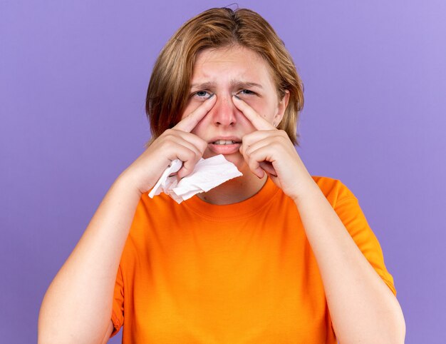 Ongezonde jonge vrouw in oranje t-shirt die zich vreselijk voelt met weefsel dat lijdt aan een loopneus, verkouden is