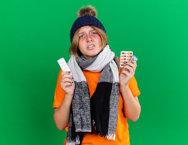 Ongezonde jonge vrouw in oranje t-shirt met hoed en warme sjaal om de nek die zich vreselijk voelt met pillen die aan griep lijden