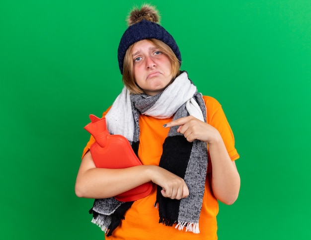Ongezonde jonge vrouw in oranje t-shirt met hoed en warme sjaal om nek voelt zich verschrikkelijk met warmwaterkruik die lijdt aan kou