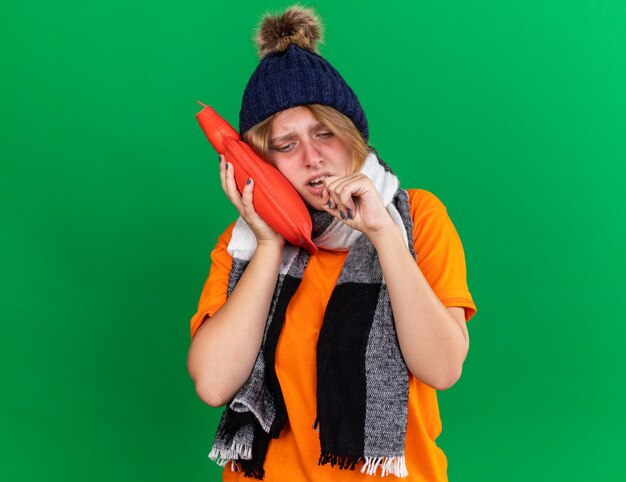 Ongezonde jonge vrouw in oranje t-shirt met hoed en warme sjaal om nek voelt zich verschrikkelijk met warmwaterkruik die lijdt aan kou