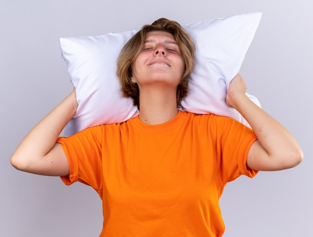 Ongezonde jonge vrouw in oranje t-shirt met kussen die zich beter voelt lachend met gesloten ogen die over een witte muur staan
