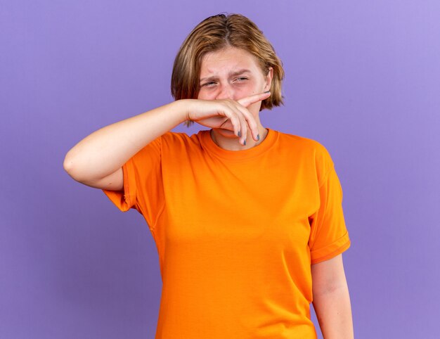 Ongezonde jonge vrouw in oranje t-shirt voelt zich verschrikkelijk en veegt haar neus af met de hand die lijdt aan een loopneus met een droevige uitdrukking die over een paarse muur staat